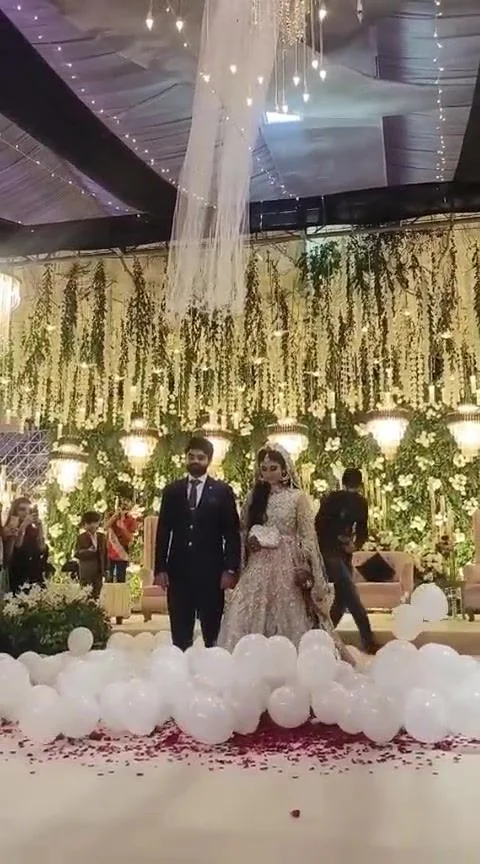 The walk-in: white balloons and petals down the aisle, chandelier florals overhead.